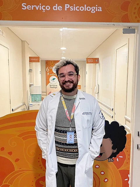 Luiz de jaleco branco sob o letreiro “Serviço de Psicologia”, no Hospital Pequeno Príncipe